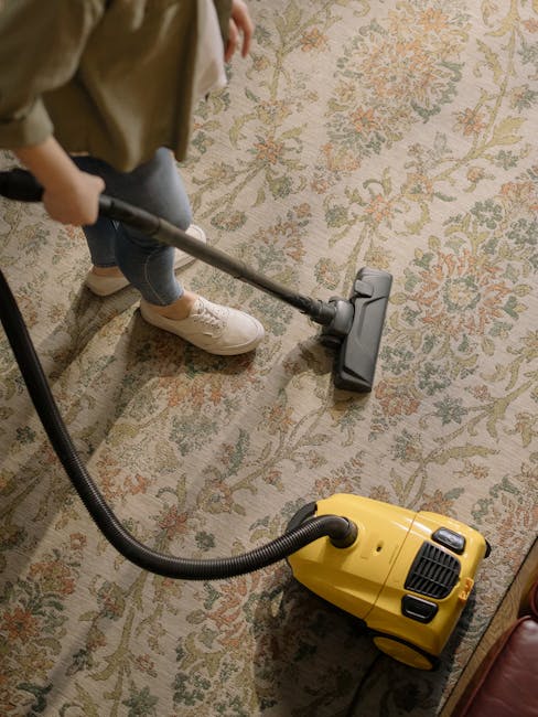 A person wearing a beige jumpsuit is performing deep cleaning on a carpeted floor in an indoor space, using a portable vacuum cleaner connected to a hose. The environment features dark and white panel