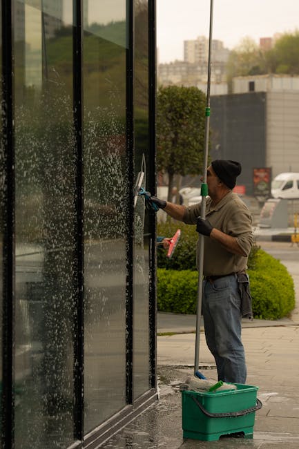 A man performing surface cleaning on a large glass window of a commercial building, using a handheld spray and squeegee, with visible soapy water and foam on the glass. The worker is dressed in casual